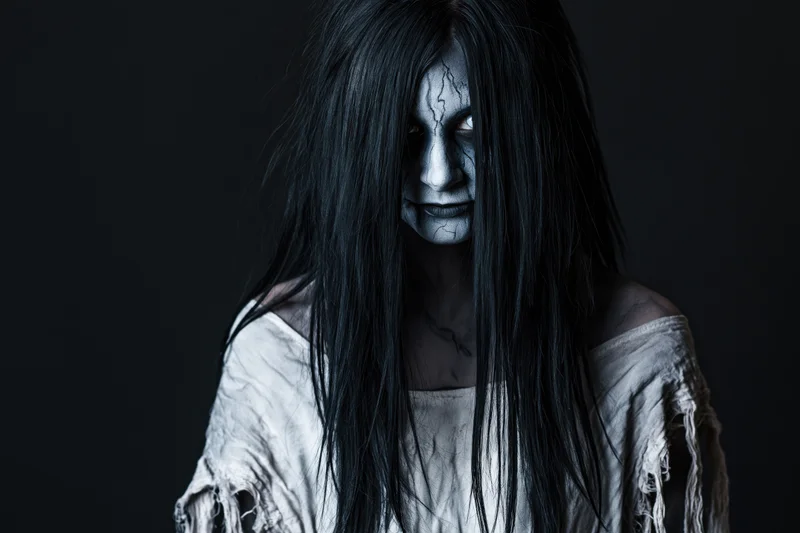 Woman in ghost makeup with cracked white face paint and long black hair, wearing tattered off-shoulder dress, studio portrait