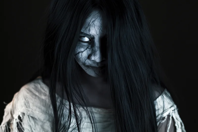 Woman with cracked white ghost makeup and long black hair in a tattered dress against dark background