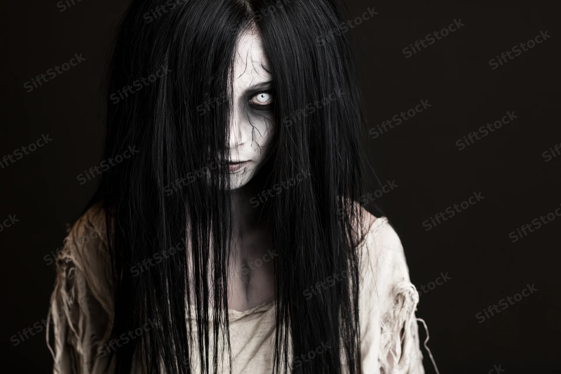 Woman with ghost makeup and long black hair in a tattered garment against a black background