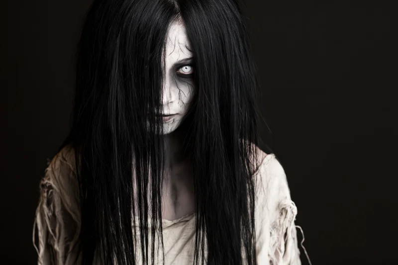 Woman with ghost makeup and long black hair in a tattered garment against a black background