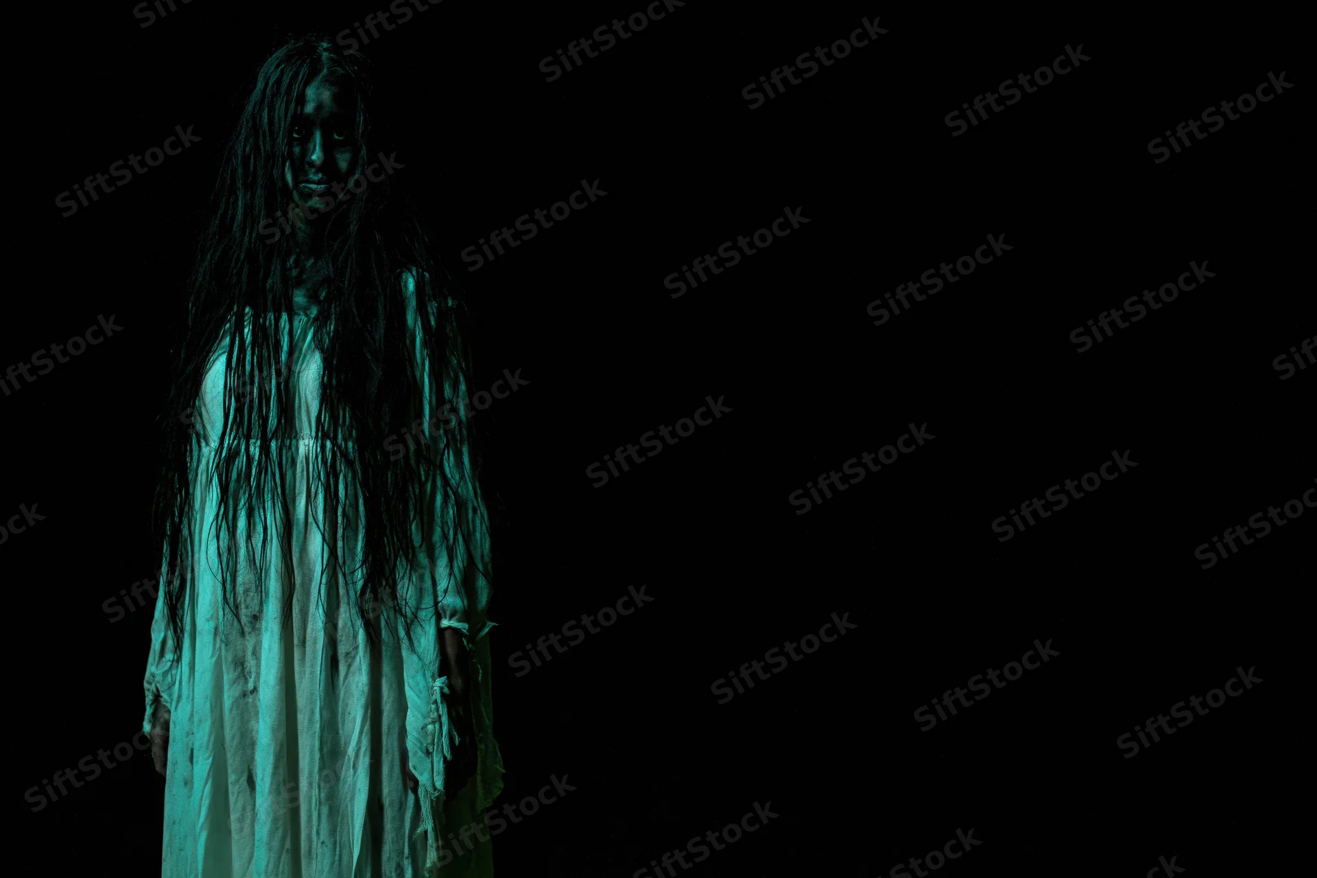 Woman in horror makeup with dark face paint and long tangled hair standing in green light against black background