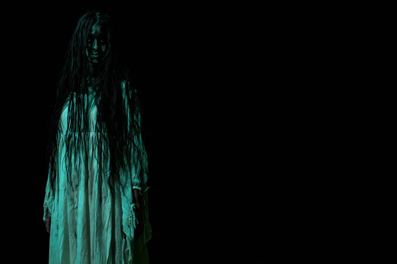 Woman in horror makeup with dark face paint and long tangled hair standing in green light against black background