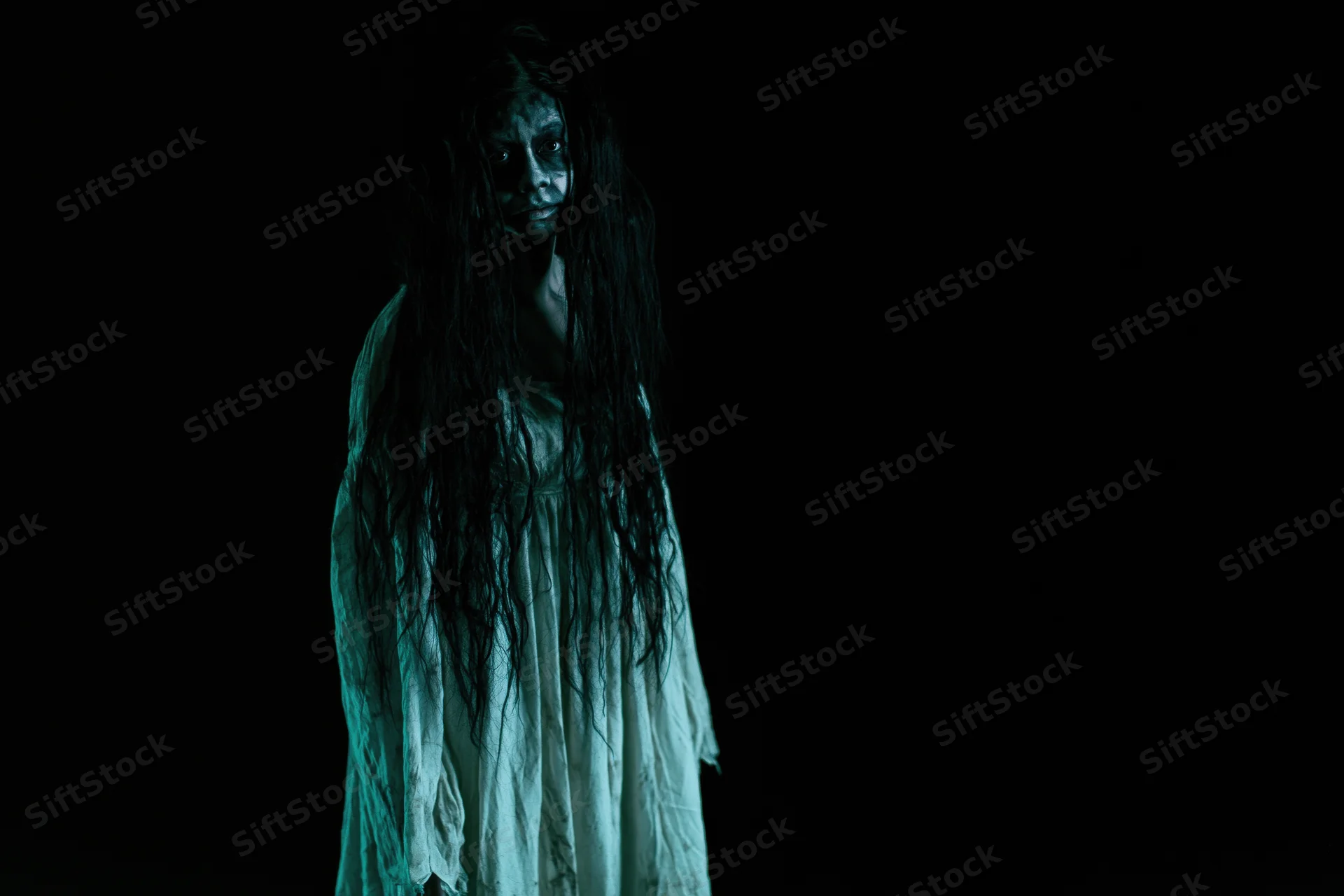 Woman in horror makeup with dark face paint and long tangled hair standing in darkness
