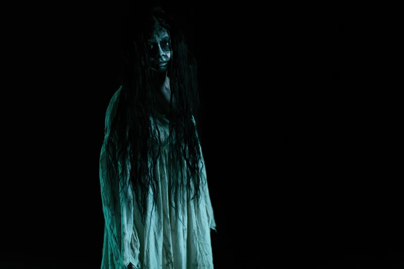 Woman in horror makeup with dark face paint and long tangled hair standing in darkness