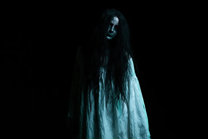 Woman in horror makeup with dark face paint and long tangled hair standing in darkness