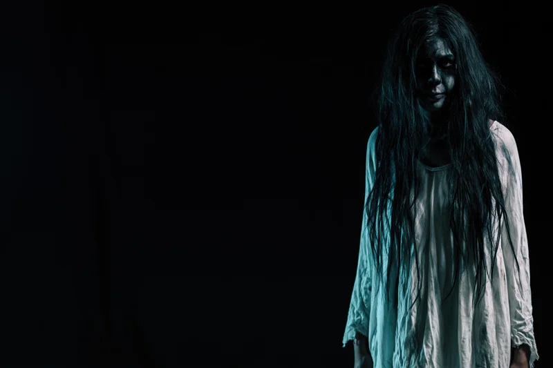 Woman in horror makeup with dark face paint and long tangled hair standing against black background
