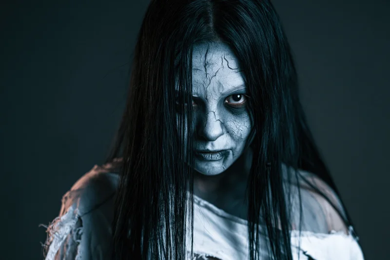 Woman with cracked grey horror makeup and long black hair in a tattered shirt, studio portrait