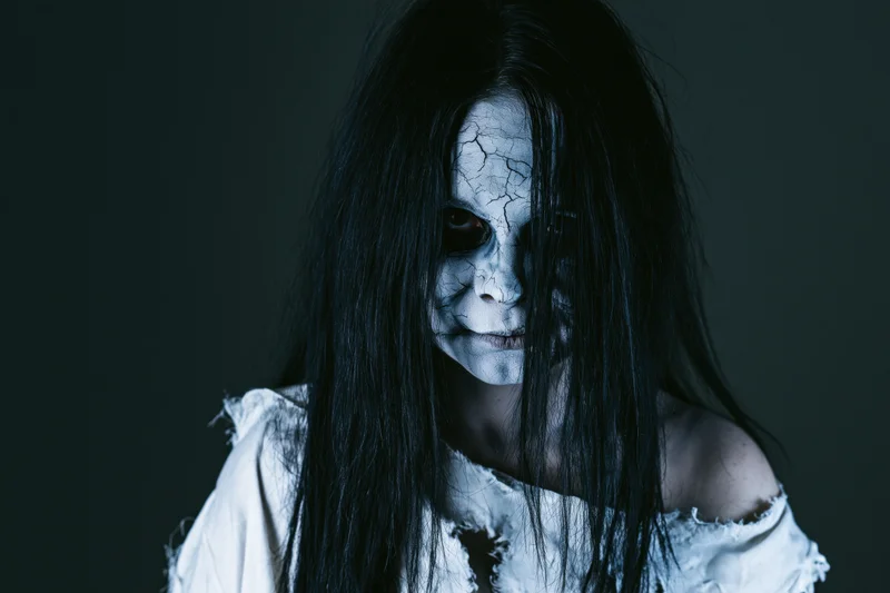 Woman with cracked pale grey face paint and long black hair in horror costume posing in dark studio