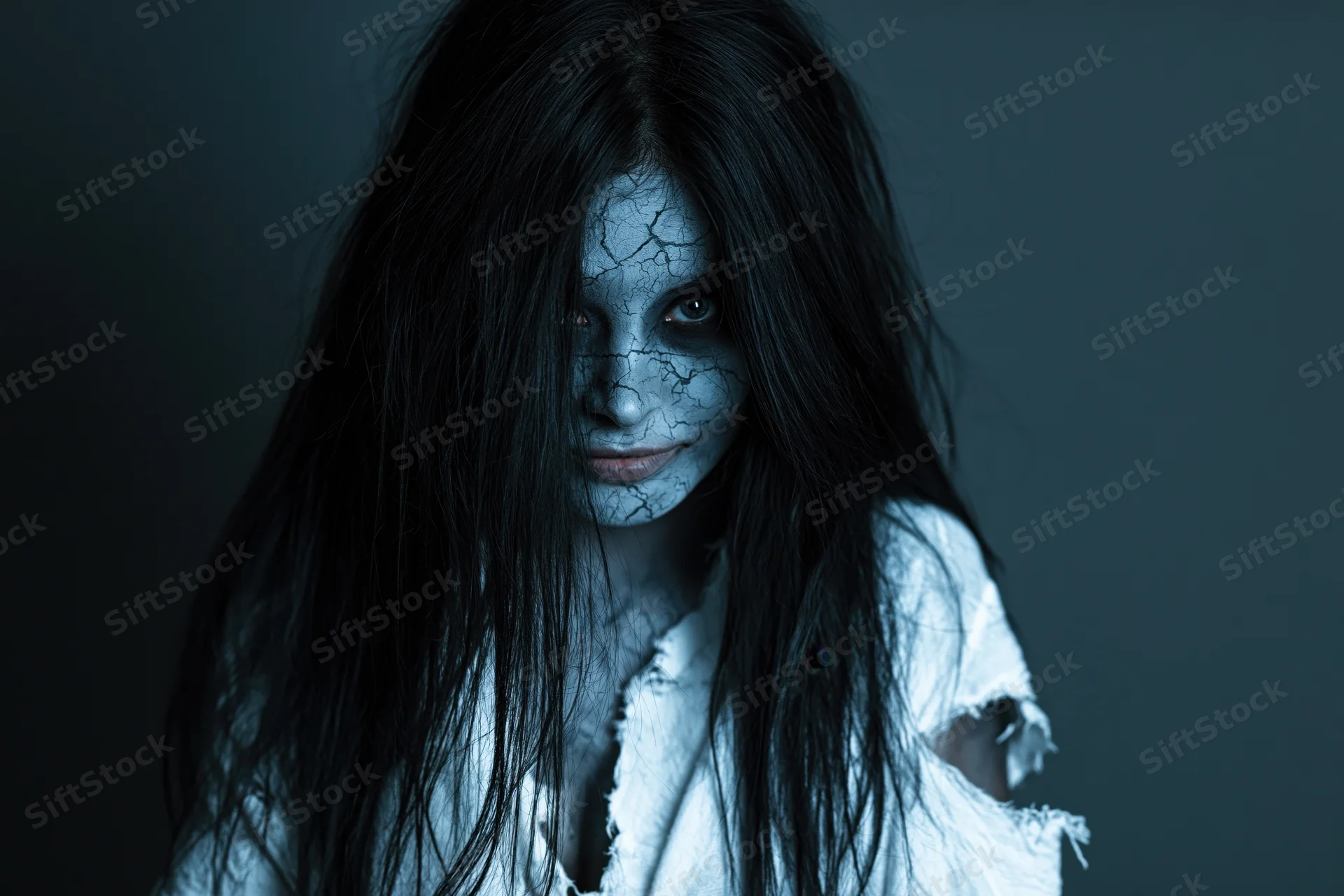 Woman with cracked pale grey face paint and long black hair in a torn white shirt against a dark background