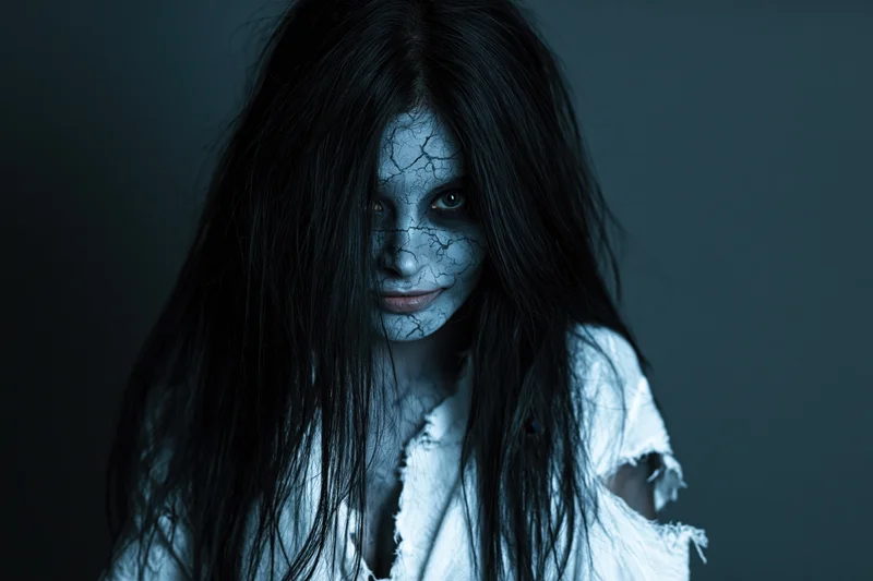 Woman with cracked pale grey face paint and long black hair in a torn white shirt against a dark background