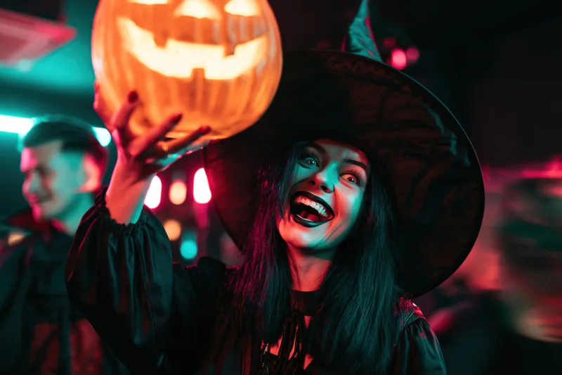 Woman in witch costume holding glowing carved jack-o-lantern at halloween party