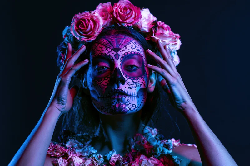 Woman with sugar skull face paint and floral crown, hands framing her face against dark background