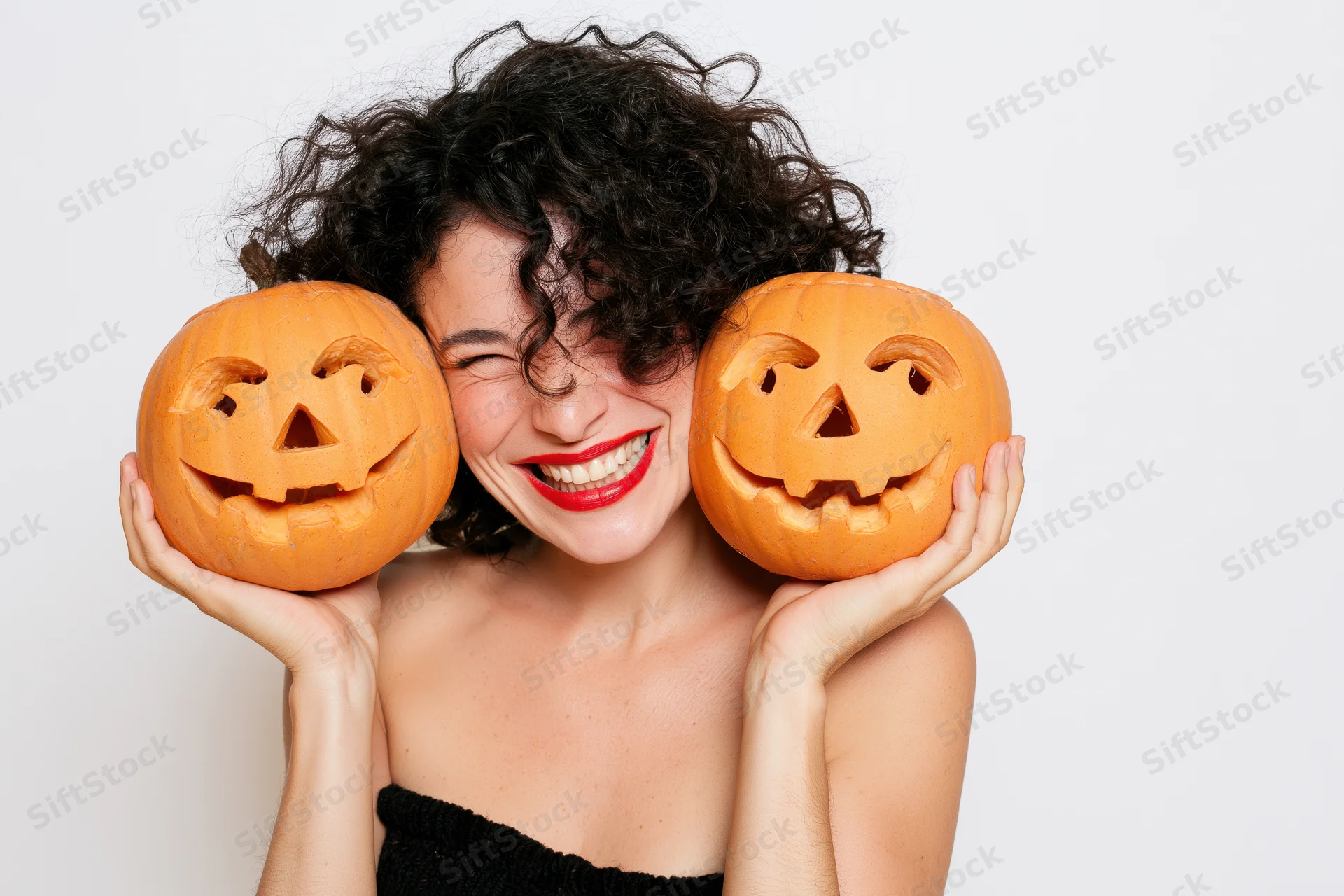 Woman winking and smiling while holding two carved jack-o-lantern pumpkins on white background