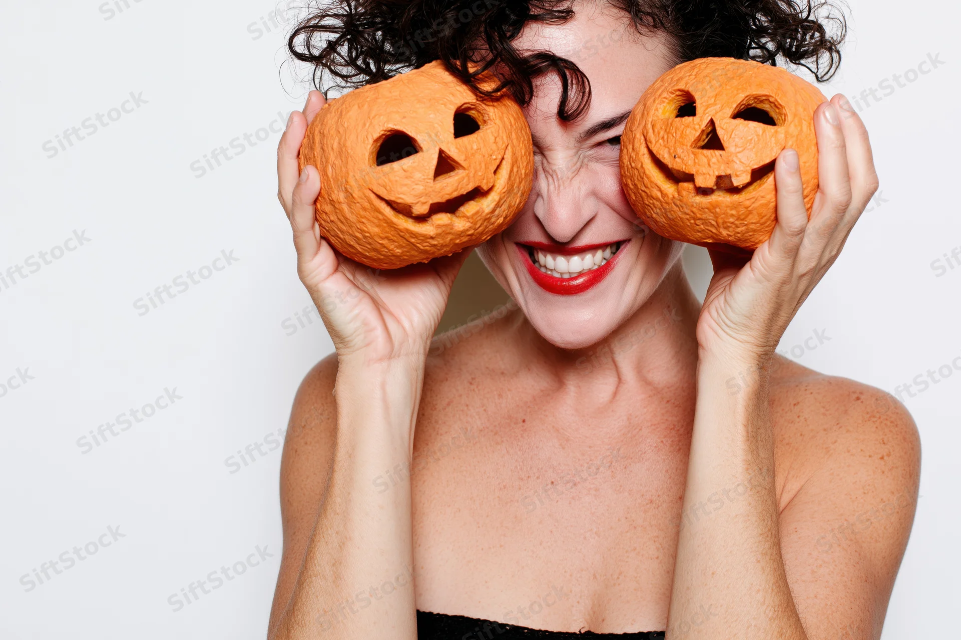 Woman holding two carved jack-o-lanterns over her eyes, smiling, in studio against white background