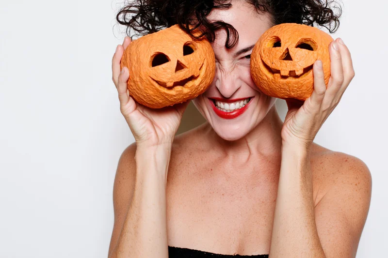 Woman holding two carved jack-o-lanterns over her eyes, smiling, in studio against white background