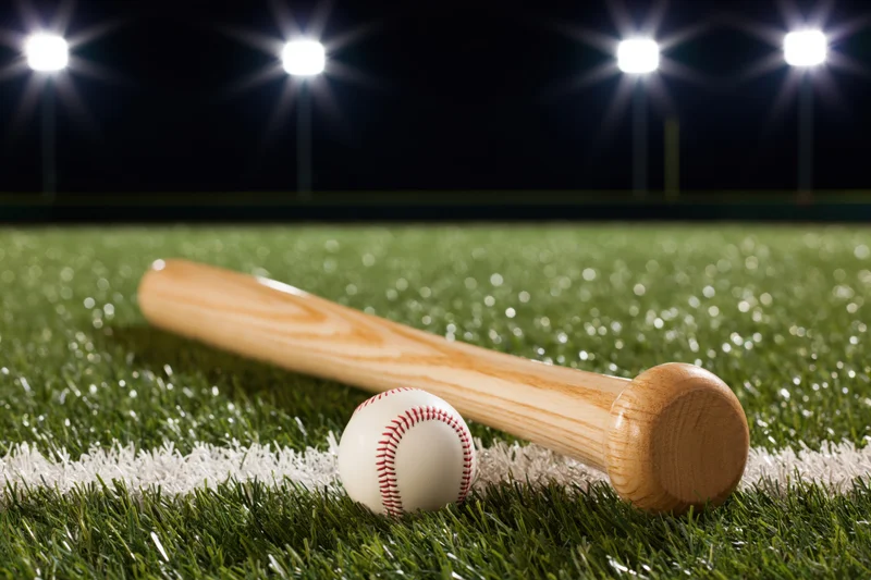 Wooden baseball bat and ball resting on green turf field under stadium lights at night