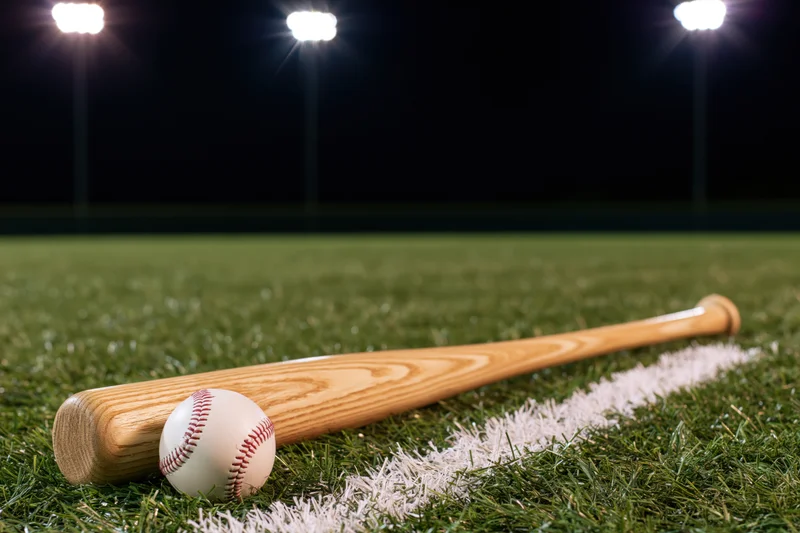 Wooden baseball bat and ball resting on grass field under stadium lights at night