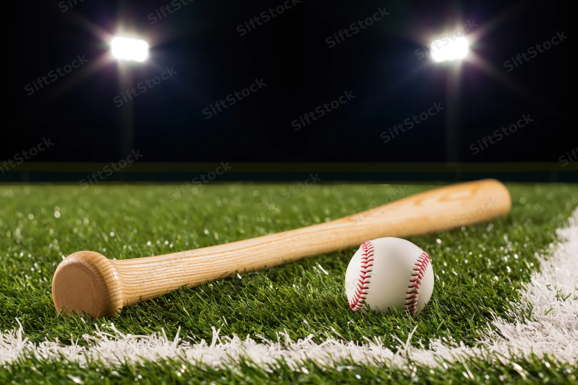 Wooden baseball bat and ball resting on green turf field under stadium lights at night