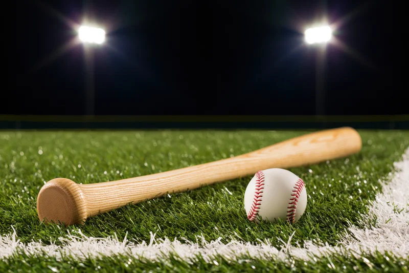 Wooden baseball bat and ball resting on green turf field under stadium lights at night