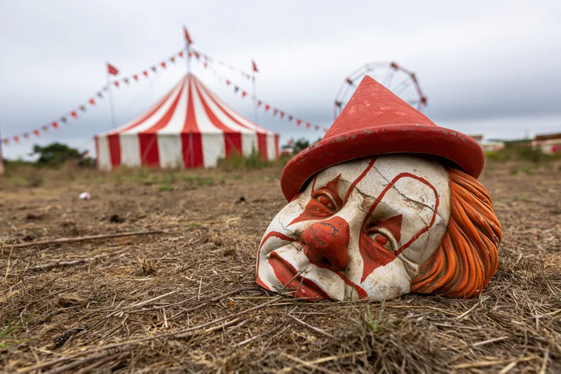 Worn ceramic clown mask with red hat and orange hair resting on dry grass in front of a striped circus tent and ferris wheel