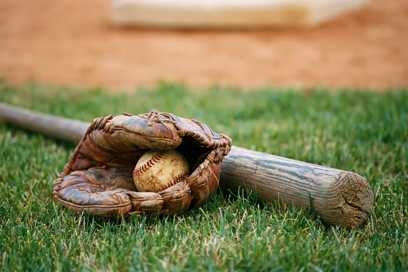 Worn leather baseball glove cradling a scuffed baseball beside a wooden bat on grass field