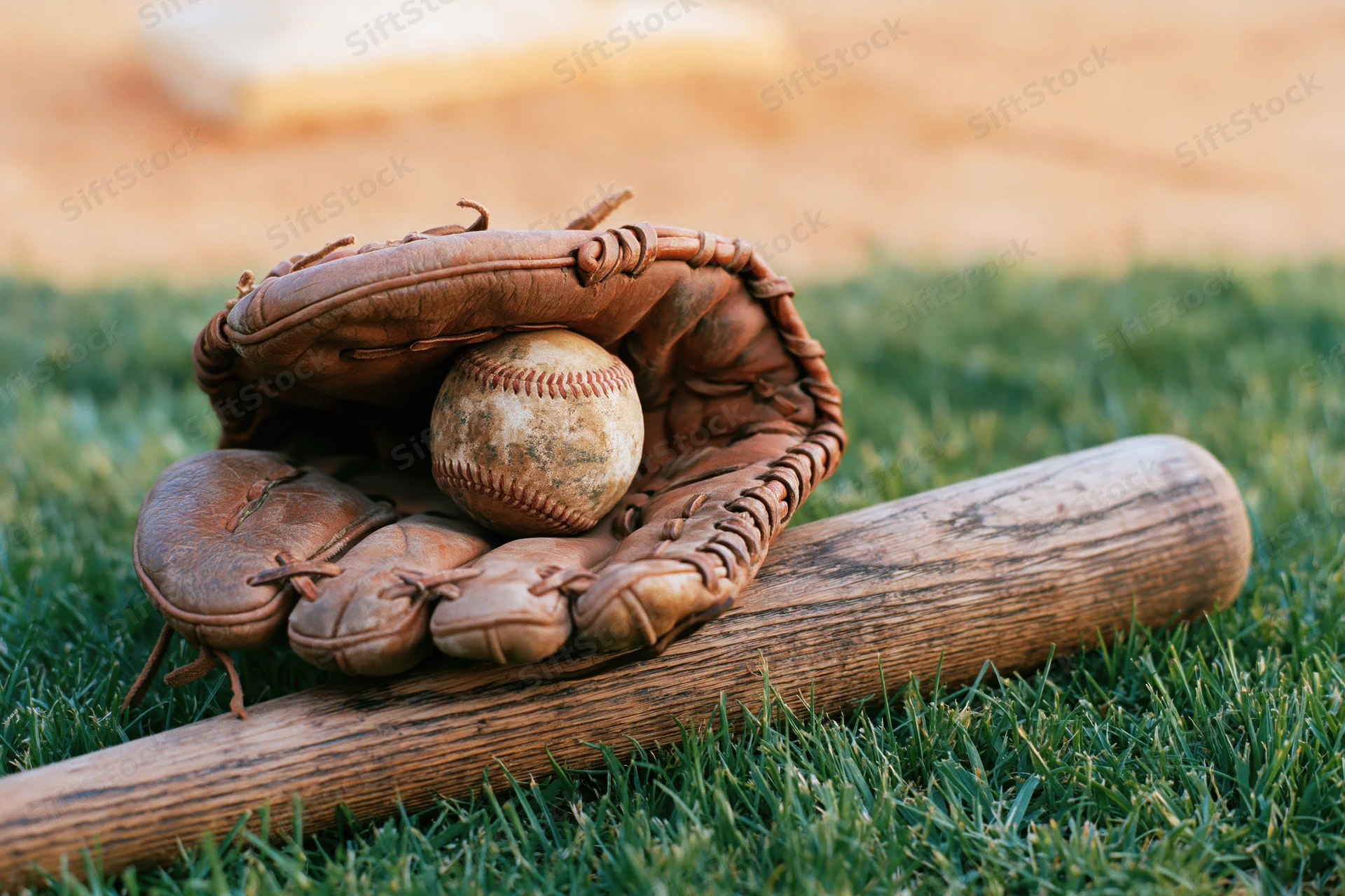 Worn leather baseball glove cradling a scuffed baseball beside a wooden bat on green grass