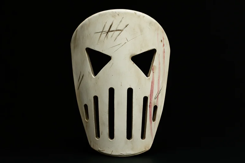 Worn white hockey mask with triangular eye cutouts and vertical slats on black background
