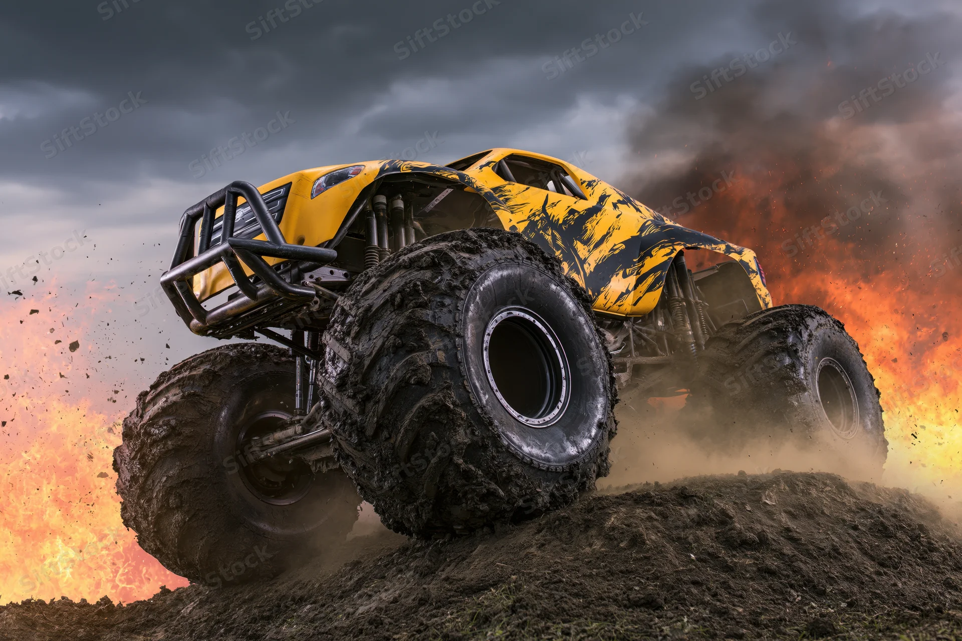 Yellow and black monster truck with oversized mud-caked tires driving over dirt hill amid fire and smoke