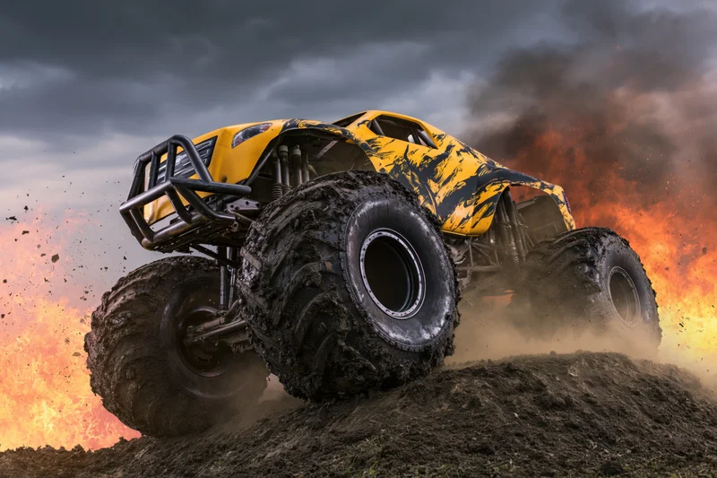 Yellow and black monster truck with oversized mud-caked tires driving over dirt hill amid fire and smoke