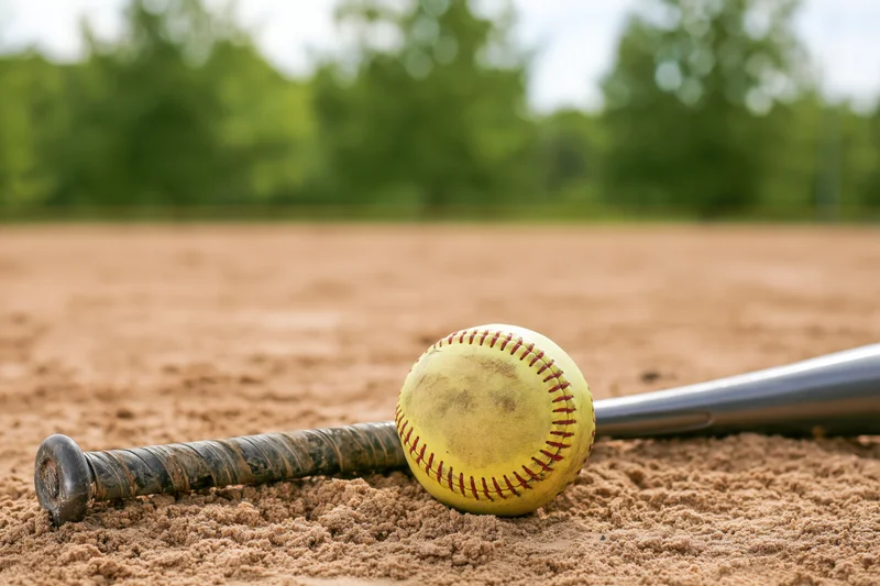 Yellow softball with red stitching resting against aluminum bat on sandy field