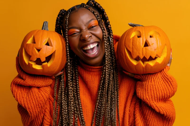 Young black woman laughing while holding two carved pumpkins against orange background