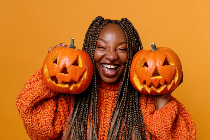 Young black woman laughing while holding two carved pumpkins in front of her face, wearing an orange knit sweater against an orange background