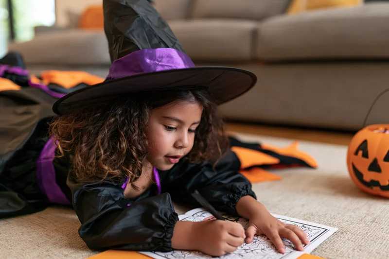 Young latina girl in witch costume coloring on carpet in living room