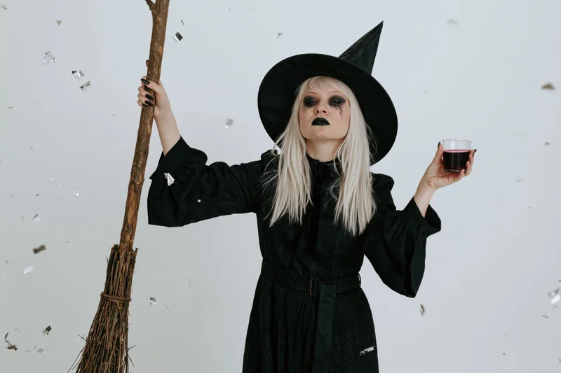 Blonde woman in witch costume holding broom and drink in studio with confetti