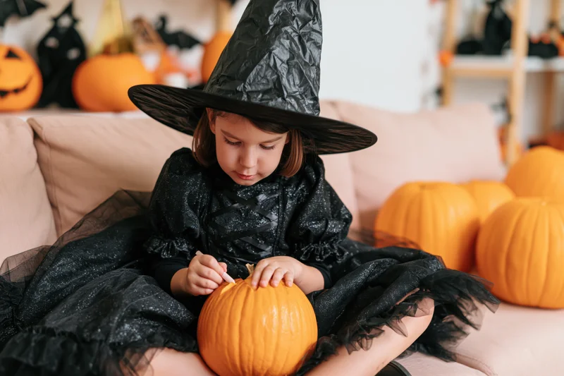 Young girl in witch costume carving a pumpkin on a sofa at home