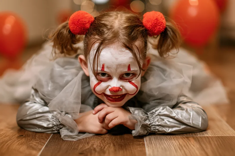 Young girl in clown costume lying on hardwood floor with face paint and red pompon pigtails