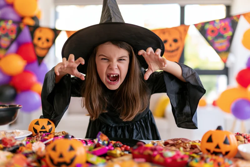 Young girl in witch costume making spooky face at candy-filled table during halloween party