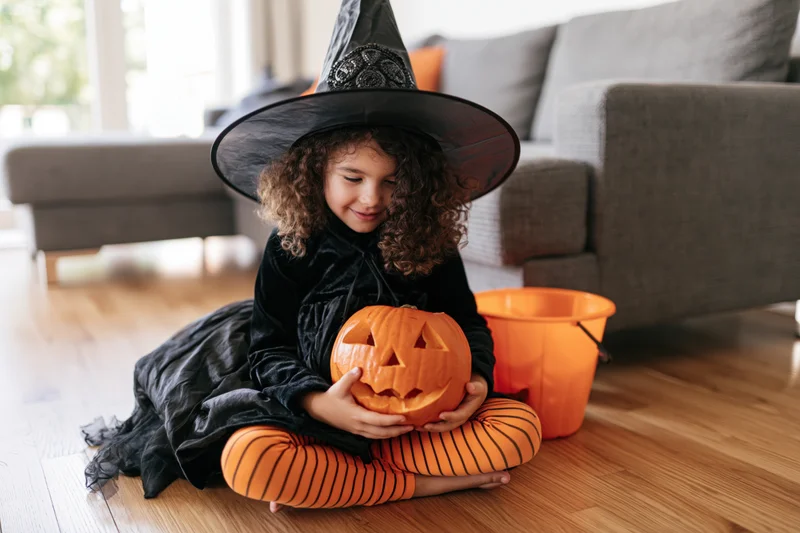Young girl in witch costume sitting on wooden floor holding carved pumpkin beside orange bucket in living room