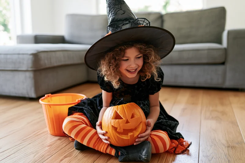 Young girl in witch costume holding carved pumpkin while sitting on wooden floor in living room
