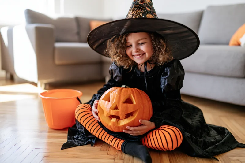 Young girl in witch costume smiling while holding carved pumpkin on wooden floor in living room