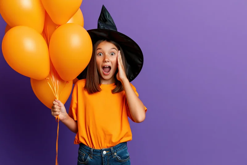 Young girl in witch hat holding orange balloons, shouting with hand to ear against purple background