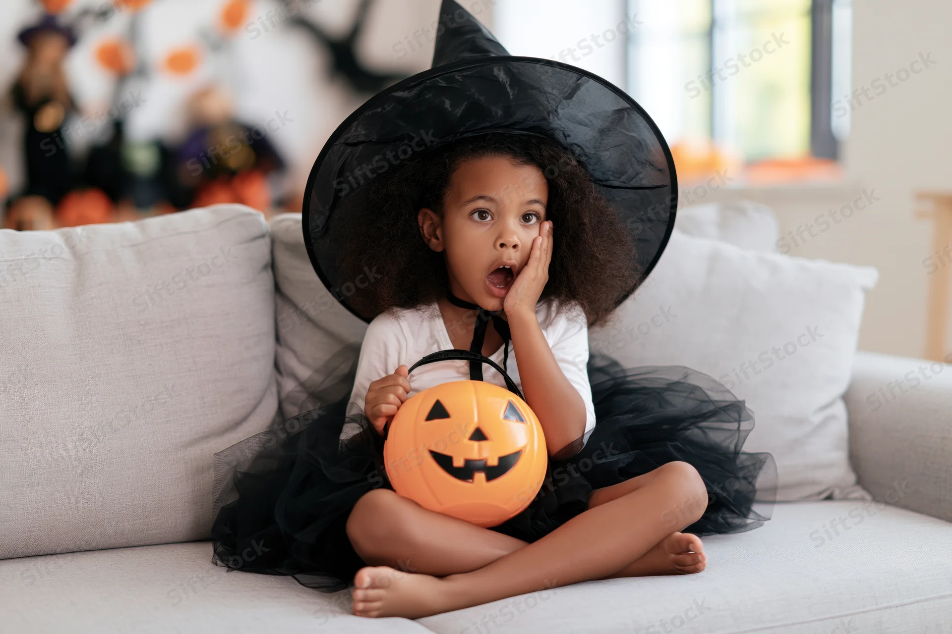 Young girl in witch costume sitting cross-legged on couch holding pumpkin bucket, surprised expression, halloween home celebration