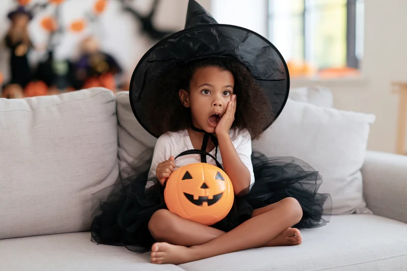 Young girl in witch costume sitting cross-legged on couch holding pumpkin bucket, surprised expression, halloween home celebration