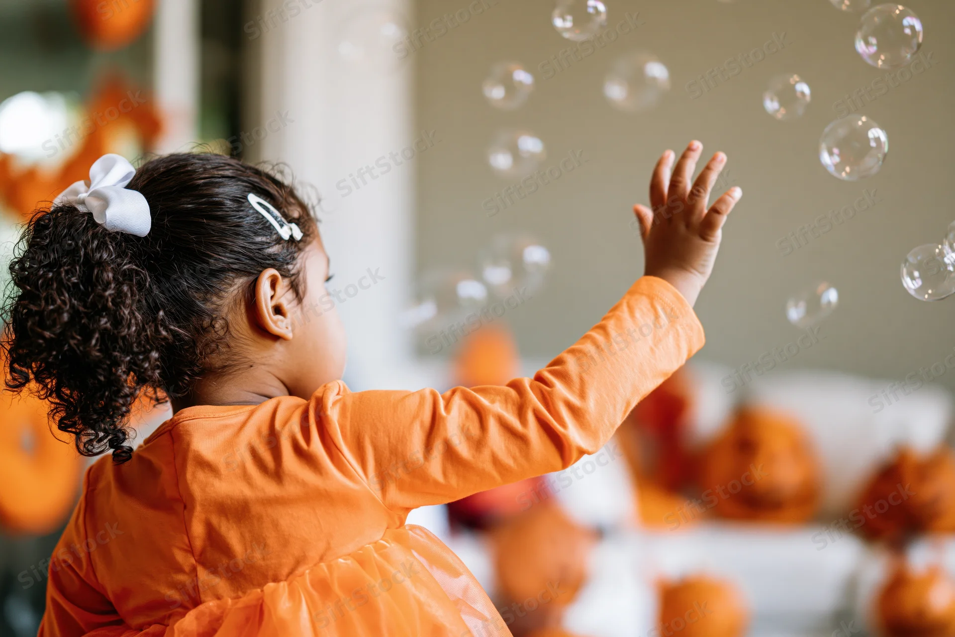 Young girl in orange halloween costume reaching for floating soap bubbles indoors