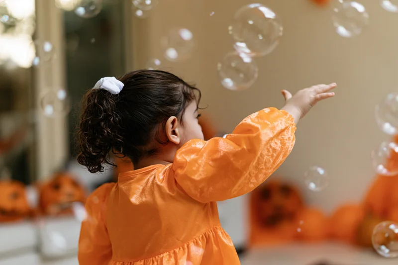 Young girl in orange halloween costume reaching for floating soap bubbles indoors