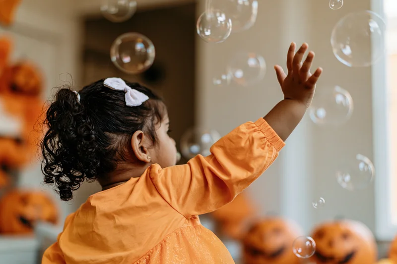 Young girl in orange halloween costume reaching for floating soap bubbles indoors