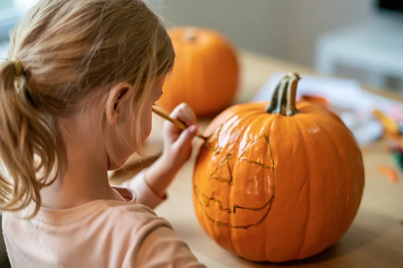 Young girl drawing a jack-o-lantern face on a pumpkin at a table indoors