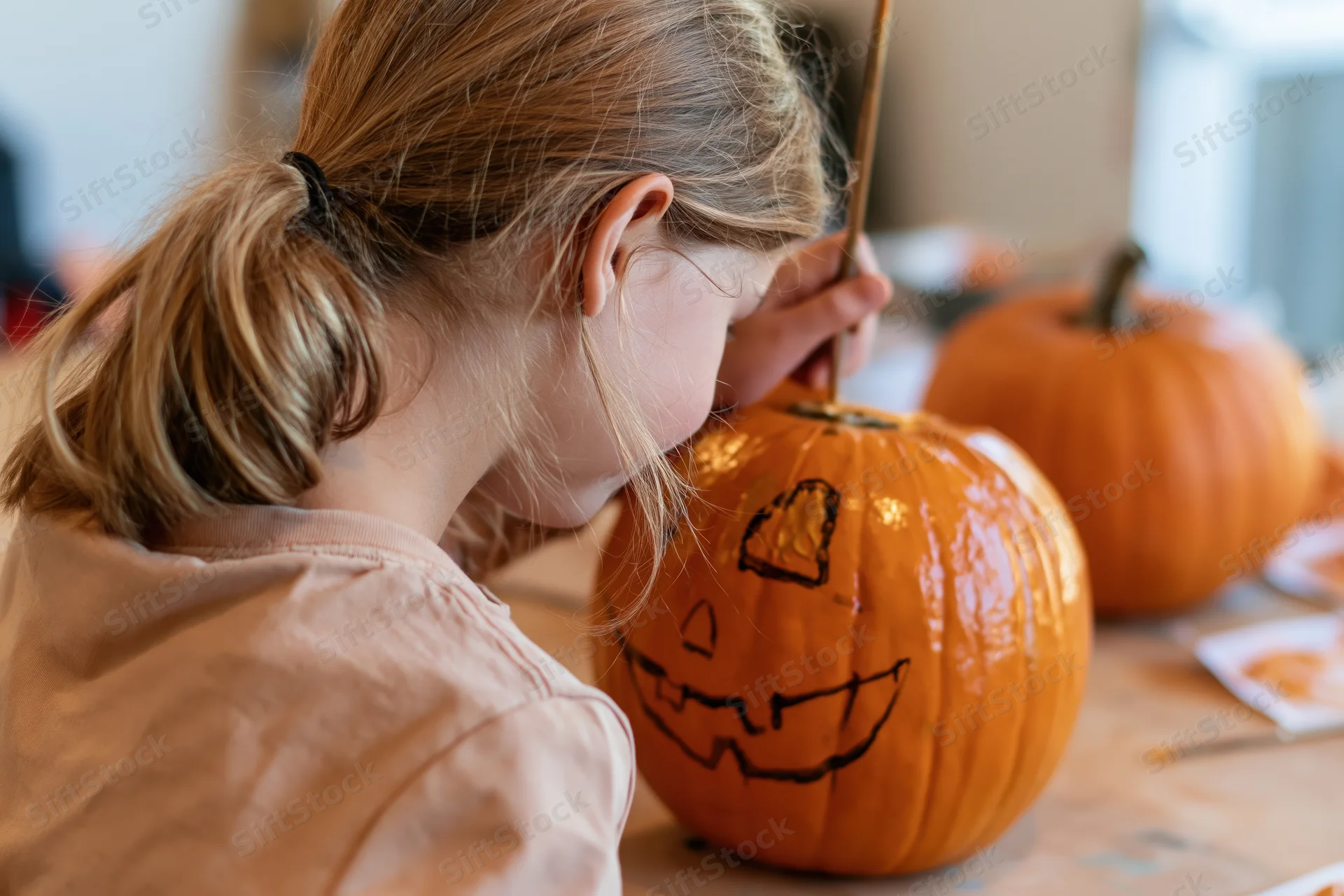 Young girl drawing a jack-o-lantern face on a pumpkin at a table indoors