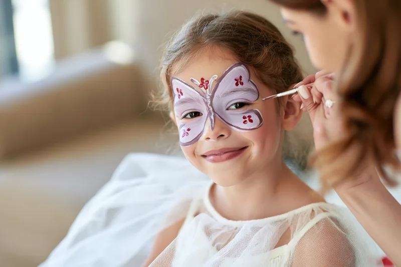 Young girl smiling as woman paints butterfly face design on her face indoors