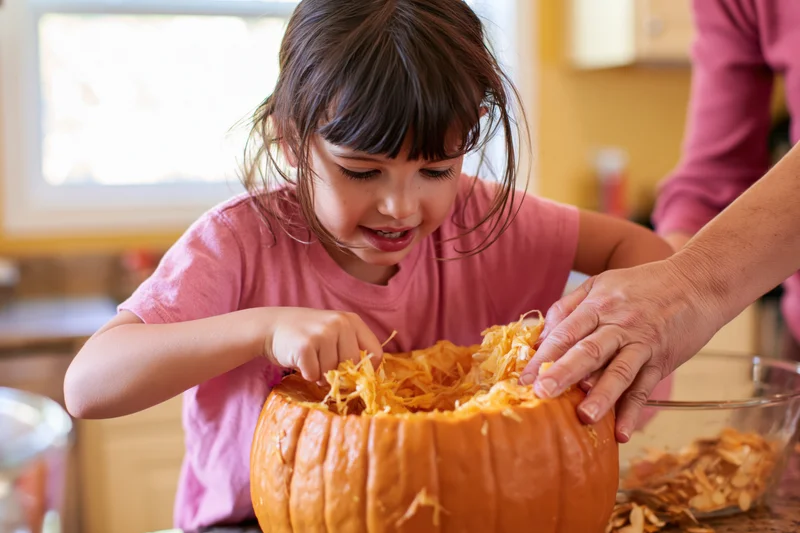 Young girl scooping stringy pumpkin guts with adult help in kitchen